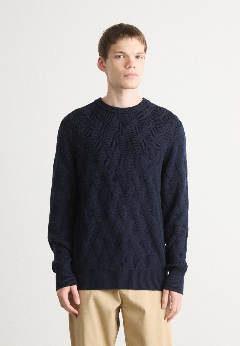 Maglione girocollo lavorato a maglia blu navy con motivo a diamante a rilievo, maniche con polsini e fondo a costine, abbinato a pantaloni beige.