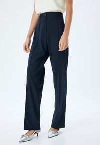 Marineblauwe high-waisted broek met een getailleerde pasvorm, voorzien van voorplooien en een enkele knoopsluiting, gecombineerd met zilveren hakken.