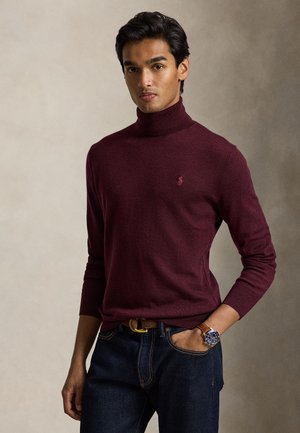 Jeune homme aux cheveux foncés portant un pull à col roulé bordeaux, un jean foncé, une montre en cuir marron et une ceinture marron, debout avec une main dans la poche.
