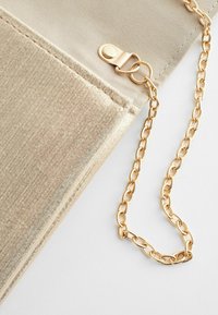 Gros plan d'un sac en tissu beige avec une bandoulière chaîne dorée attachée par un fermoir doré sur fond blanc.