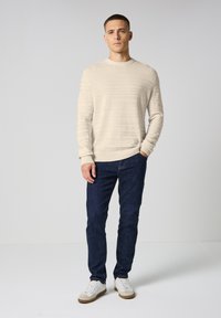 Pull en maille côtelée beige clair avec un col rond, associé à un jean slim bleu foncé et des baskets blanches avec des accents beiges.