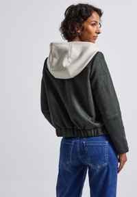 Vrouw met kort, krullend haar, gekleed in een zwarte jas met een beige capuchon en blauwe spijkerbroek, staand en kijkend opzij.