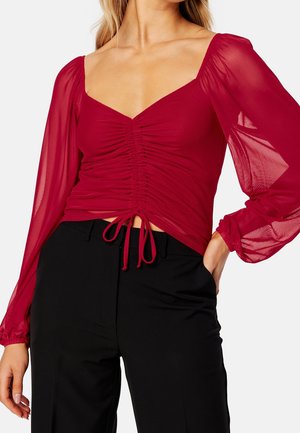 Femme portant un haut rouge ajusté à manches longues froncées avec des manches bouffantes transparentes et un pantalon noir taille haute, main dans la poche.