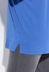T-shirt in cotone blu con vestibilità rilassata, caratterizzato da una fessura laterale all'orlo. Abbinato a pantaloni in tessuto grigio aderente, che mostrano una texture liscia.