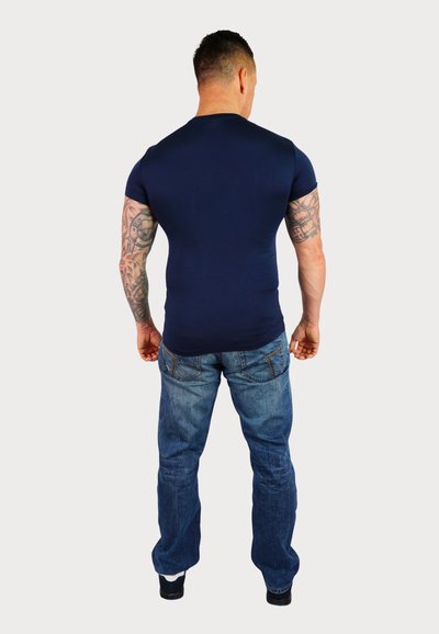 Navyblaues, körperbetontes T-Shirt aus dehnbarem Material, kombiniert mit hellblauen Jeans. Sichtbare Tattoos an den Armen, stehend vor einem weißen Hintergrund.