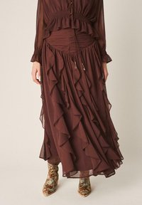 Jupe maxi marron à volants superposés associée à des bottines pointues à motif serpent, portée avec un haut assorti marron plissé avec des boutons.