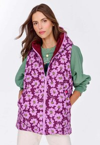 Kobieta wpurpurowej kwiecistej kamizelce puchowej z kapturem, nosząca zieloną bluzę, stoi z rękami w kieszeniach kamizelki na tle białym.