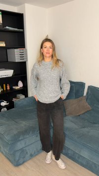 Άνετο γκρι πλεκτό πουλόβερ συνδυασμένο με μαύρο παντελόνι φαρδιάς γραμμής και λευκά αθλητικά παπούτσια. Το δωμάτιο έχει έναν υφασμάτινο μπλε καναπέ και μια σκούρα βιβλιοθήκη.