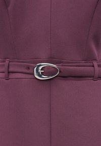 Tumši sarkans kleita ar gludu audumu, jostu viduklī un sudraba ovālu sprādzi. Tai ir tīras līnijas un minimālisma dizaina detaļas.