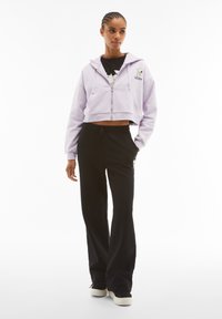 Felpa corta di lavanda con zip frontale e grafica di Snoopy, abbinata a pantaloni della tuta neri con dettagli di teschio bianchi. Texture liscia.