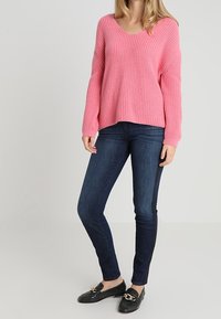 Femme portant un pull en tricot rose ample, un jean skinny bleu foncé et des mocassins noirs avec des accents en chaîne dorée, se tenant devant un fond clair.
