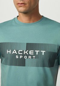 Muž v modrozeleném tričku s černými vertikálními pruhy a bílým nápisem "HACKETT SPORT" přes hruď, střižené na krku a ramenou.