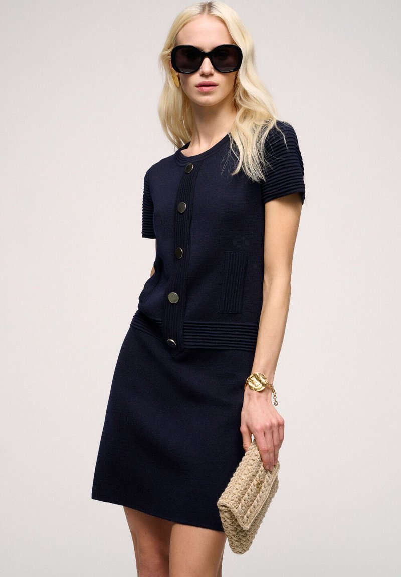 Abito lavorato a maglia blu navy con maniche corte, texture a coste, scollo rotondo e cinque bottoni. Accoppiato con una clutch intrecciata beige.