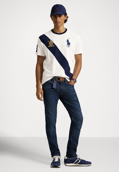 Λευκό κοντομάνικο T-shirt με ναυτικό ρίγα και λογότυπο, συνδυασμένο με σκούροτζιν και μπλε αθλητικά παπούτσια. Περιλαμβάνει ναυτικό καπέλο και ζώνη με ρίγες.