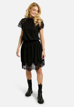 Žena s dlhými blond vlasmi v čiernych šatách s čipkovanými rukávmi a lemom, v čiernych členkových topánkach, usmievajúca sa a pozerajúca nadol.