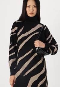 Schwarzer Strickpullover mit beigen Zebra-Streifen, mit einem hohen Kragen und langen Puffärmeln. Das Model hält eine kleine schwarze Handtasche.