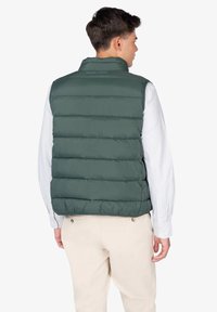 Chaleco acolchado verde con costuras horizontales, cuello alto y logo en la parte posterior, llevado sobre una camisa a rayas clara y pantalones caqui.