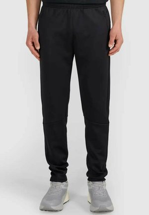 Mężczyzna w czarnych zwężanych spodniach dresowych i szarych sportowych butach, stojący na jednolitym szarym tle.