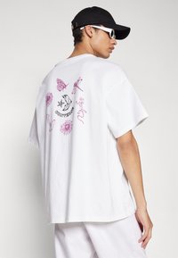 Tricou alb din bumbac, cu o croială lejeră. Prezintă desene grafice roz cu insecte și flori pe spate, și textul "Converse" în centru.