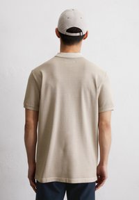 Beżowa koszulka polo o teksturowanej tkaninie, z krótkimi rękawami i płaskim kołnierzykiem, zestawiona z dopasowaną jasną czapką. Widok tylko z tyłu.