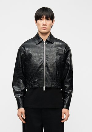 Joven con chaqueta corta negra de cuero sintético con textura de cocodrilo, cremallera frontal y cinturilla elástica, posando frente a un fondo liso.