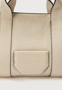 Detalhe de alças de bolsa de couro texturizado bege e bolso frontal com nome da marca sutilmente em relevo.