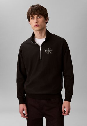 Joven con una sudadera negra Calvin Klein con cremallera hasta un cuarto sobre una camisa blanca, de pie frente a un fondo gris liso.