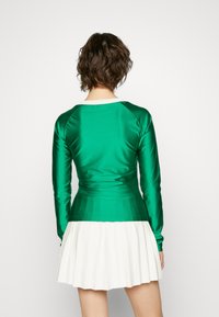 Top de satén verde de manga larga con un diseño ajustado, combinado con una falda blanca de pliegues, que presenta una textura horizontal y colores contrastantes.