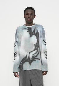 Πουλόβερ με μακριά μανίκια σε διαβάθμιση μπλε και λευκού, με μαύρα φυλετικά γραφικά στα μανίκια και το σώμα. Το υλικό φαίνεται υφασμάτινο και πλεκτό.