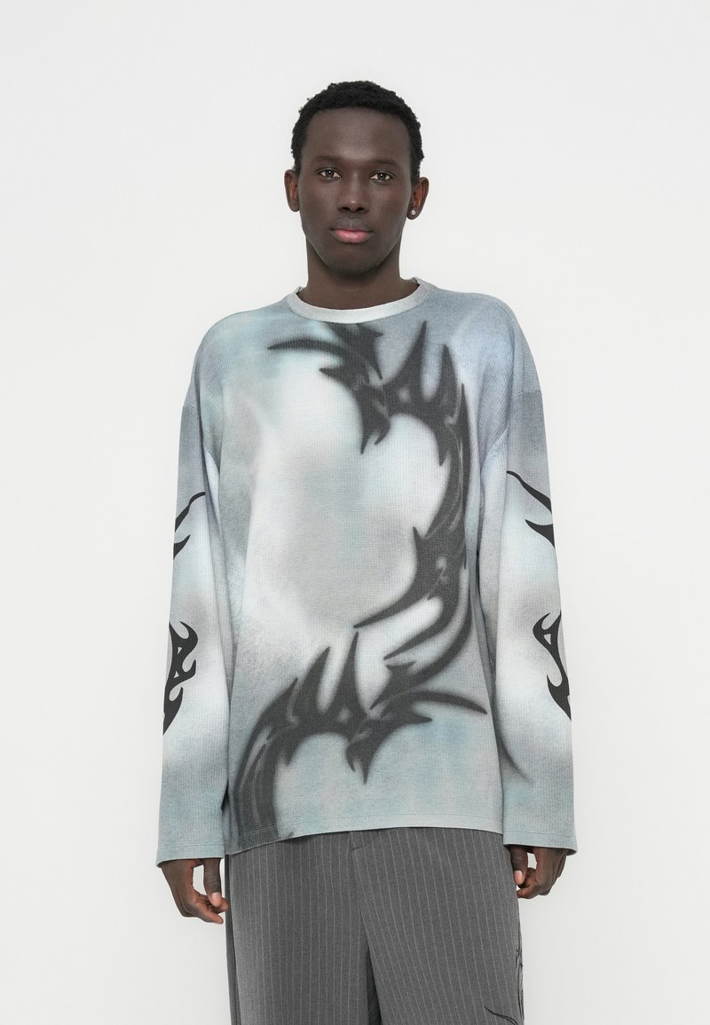 Πουλόβερ με μακριά μανίκια σε διαβάθμιση μπλε και λευκού, με μαύρα φυλετικά γραφικά στα μανίκια και το σώμα. Το υλικό φαίνεται υφασμάτινο και πλεκτό.