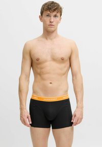 Melnas bokseru apakšbikses ar oranžu jostu, kurā ir rakstīts 'JACK & JONES'. Audums izskatās gluds un pieguļošs, uzsverot formas. 