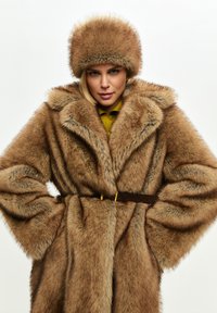 Cappotto in pelliccia sintetica marrone con ampio colletto, vita aderente e finitura testurizzata. Accompagnato da un cappello di pelliccia marrone coordinato.