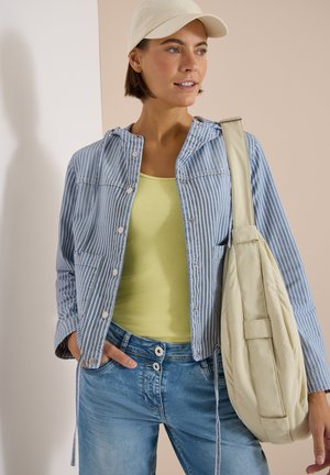 Femme portant une casquette beige, une veste boutonnée à rayures bleues et blanches par-dessus un haut jaune, un jean en denim, portant un grand sac à épaule beige.