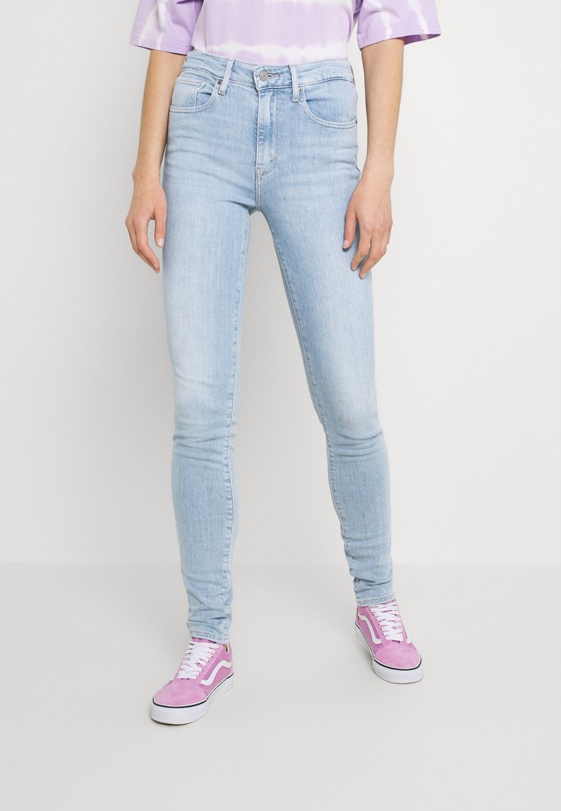 Personne portant un jean skinny bleu clair, un t-shirt violet tie and dye et des baskets roses, debout devant un fond blanc uni.