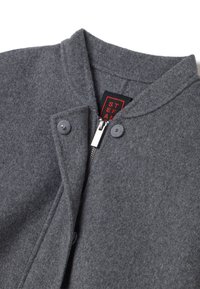 Veste en laine grise avec un col haut, des boutons sur le devant et une fermeture éclair en métal. Étiquette visible en rouge et noir à l'intérieur du col.