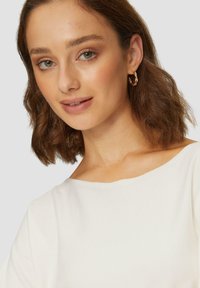 Haut blanc à côtes avec un large col, présentant un mannequin avec des cheveux ondulés jusqu'aux épaules et des boucles d'oreilles en torsade dorées.
