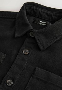 Schwarzes Hemd aus weichem Stoff mit Button-Down-Kragen, ausgestattet mit zwei Brusttaschen und braunen Kunststoffknöpfen. Das Etikett lautet "NEXT."