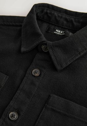 Juodas marškiniai iš minkštos medžiagos su mygtukų apykakle, turintys du priekiniai kišeniai ir rudos plastikinės mygtukai. Etiketė sako "NEXT."