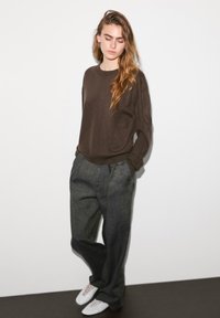 Pull en laine marron associé à un pantalon gris ample et des sneakers blanches. La tenue présente des lignes simples et une coupe décontractée. Design minimaliste.