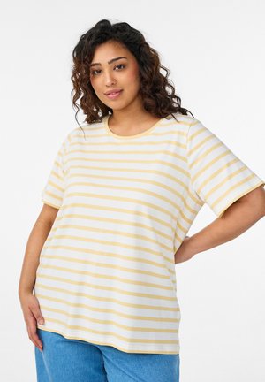 Frau mit lockigem Haar, die ein gelb-weiß gestreiftes T-Shirt und blaue Jeans trägt, posiert mit einer Hand auf der Hüfte vor einem weißen Hintergrund.