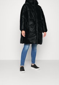 Cappotto imbottito nero con cappuccio, dotato di chiusura con zip e due tasche laterali. Indossato sopra jeans blu e scarpe slip-on nere.