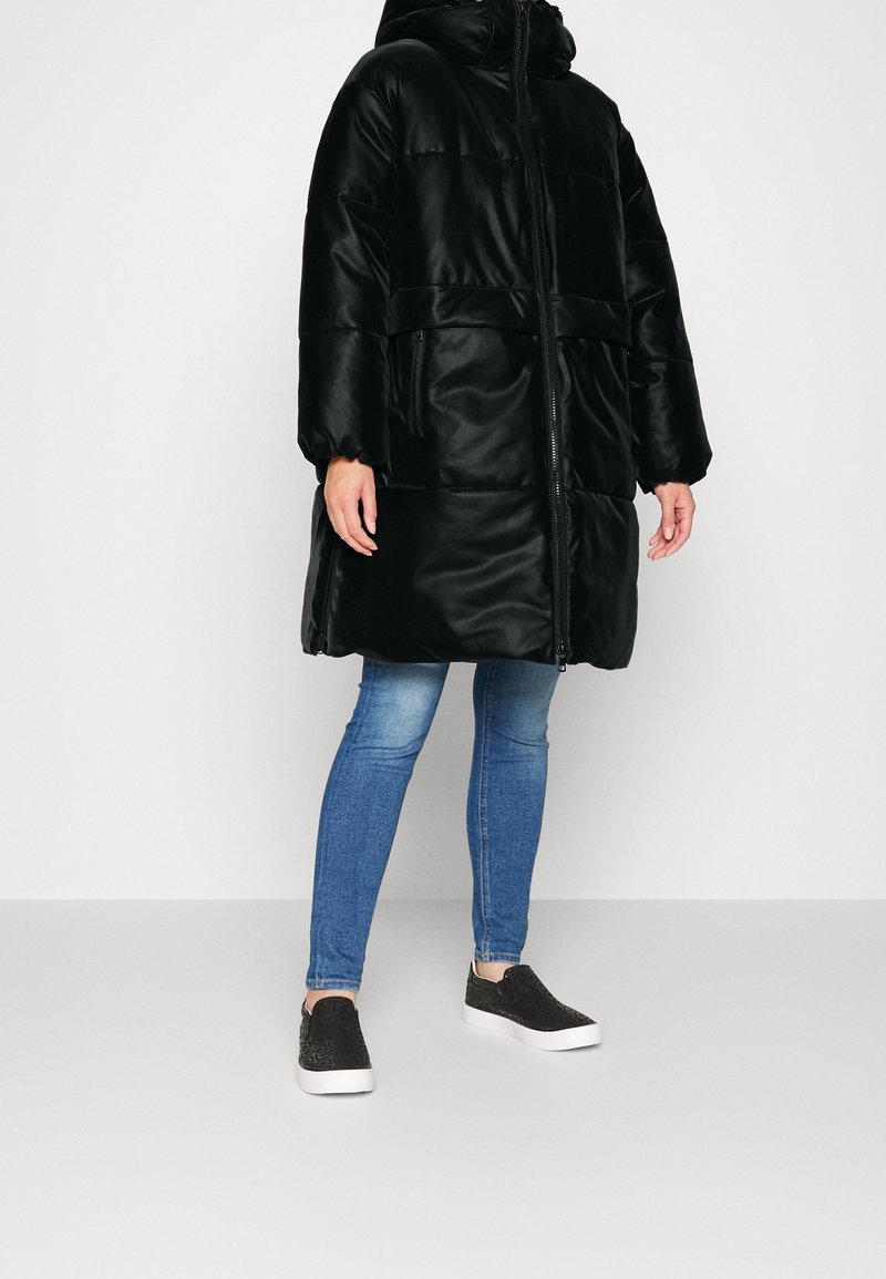 Cappotto imbottito nero con cappuccio, dotato di chiusura con zip e due tasche laterali. Indossato sopra jeans blu e scarpe slip-on nere.