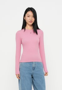Long-sleeve pink ribbed top with a round neckline and a small logo at the chest, paired with light blue loose-fit denim jeans.