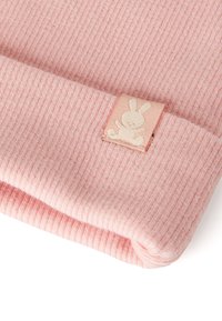 Gorro de punto acanalado de color rosa suave con puño doblado que presenta una etiqueta con un logo de conejito blanco. La textura parece cálida y acogedora.