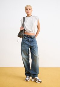 Valkoinen lyhythihainen urheilupaita, cropped-malli, vaaleansiniset löysät farkut haalistuneilla alueilla, musta vyö metallikoristeilla ja urheilukengät.