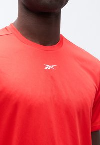 Άνδρας που φορά φωτεινό κόκκινο αθλητικό μπλουζάκι με μικρό λευκό λογότυπο Reebok στο κέντρο του στήθους, πάνω σε απλό ανοιχτό φόντο.