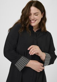 Glimlachende vrouw met schouderlang bruin haar, gekleed in een zwarte blouse met lange mouwen en versierde manchetten, die met haar handen één manchette aanpast.