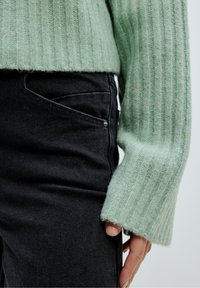 Světle zelený žebrovaný pletený svetr s širokým manžetovým rukávem, spárovaný s černými džíny s přední kapsou. Textura vypadá měkce a útulně.