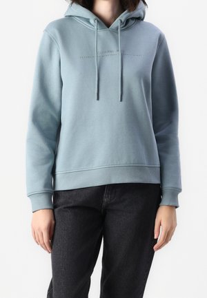 Persoon draagt een lichtblauwe hoodie met voorzak en trekkoorden, gecombineerd met een zwarte broek tegen een effen achtergrond.