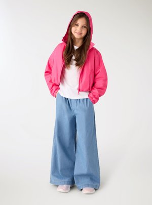 Ragazza con lunghi capelli castani che indossa una giacca rosa con cappuccio, una camicia bianca, jeans blu a gamba larga e sneakers rosa chiaro, in piedi con le mani nelle tasche.